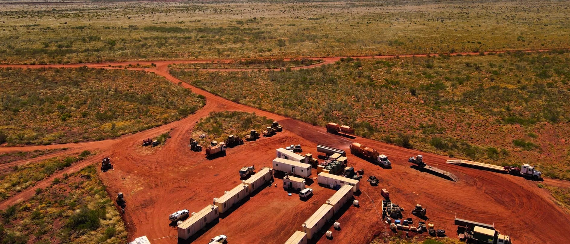 Aerial photograph of a remote camp and laydown yard of transportable buildings, trucks and plant on red dirt, surrounded by scrub