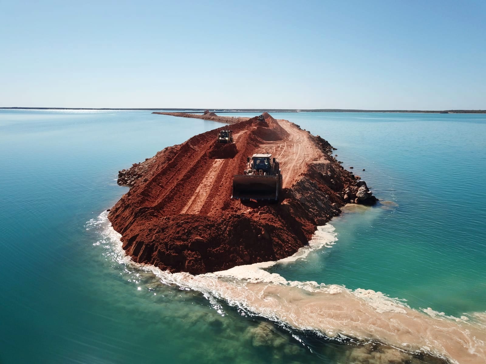 Aerial photograph of a wall of red fill pushed out into turquoise water at Useless Loop, with a wheel loader and an excavator working on top of it