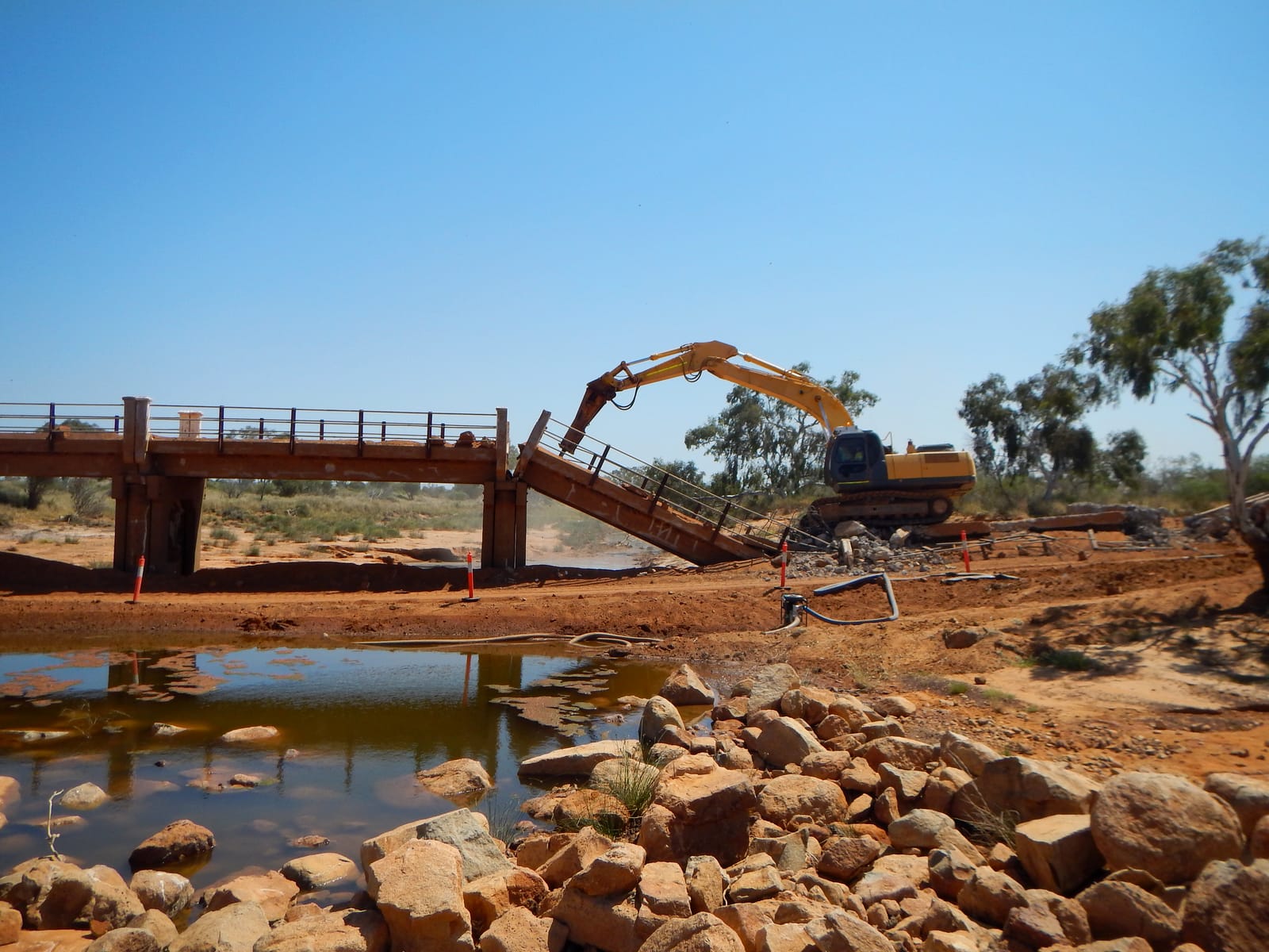 An excavator working beside a timber bridge over a Murchison river pool, with fresh rock armouring stacked along the bank in the foreground