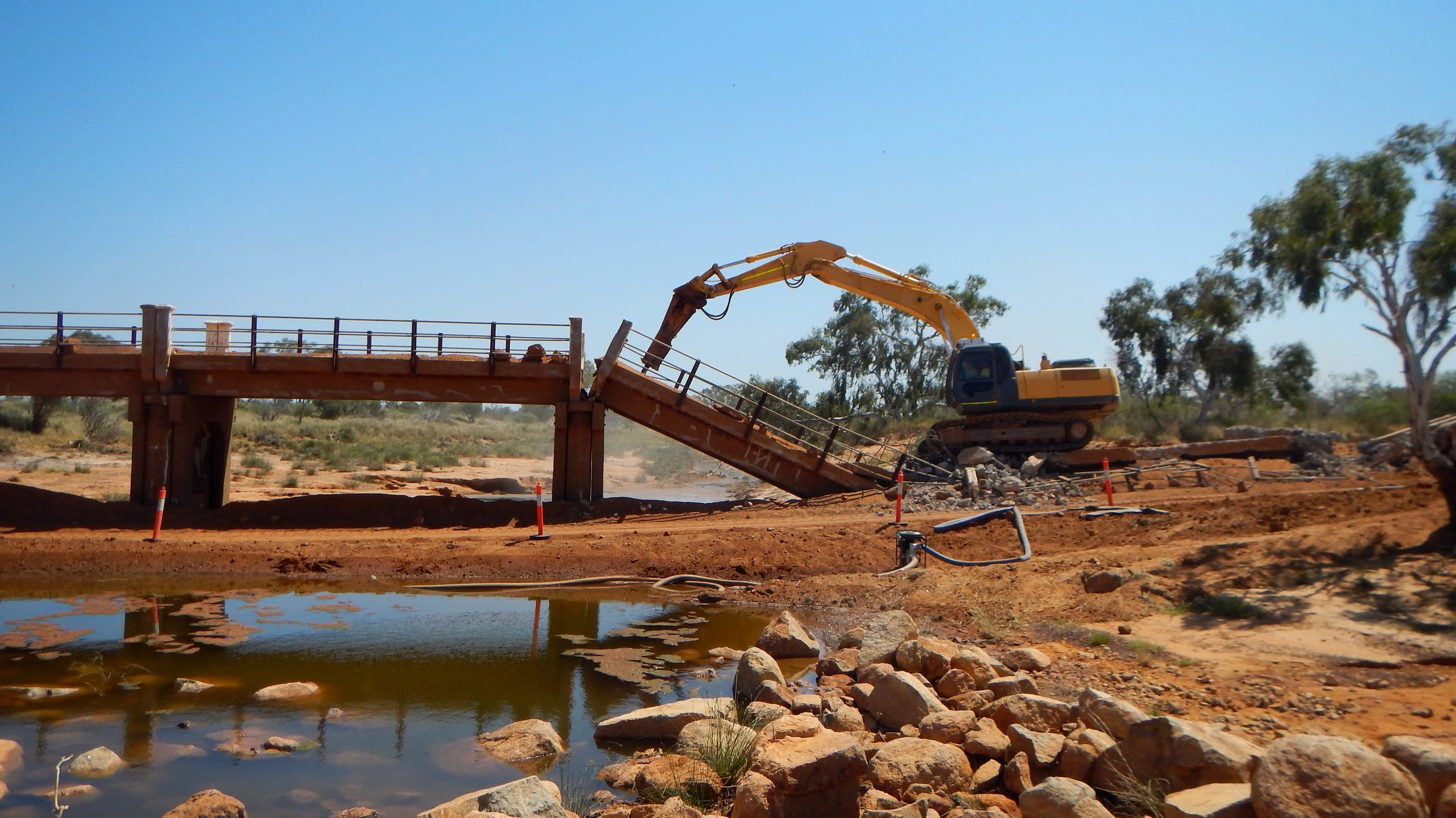 An excavator working beside a timber bridge over a Murchison river pool, with fresh rock armouring stacked along the bank in the foreground