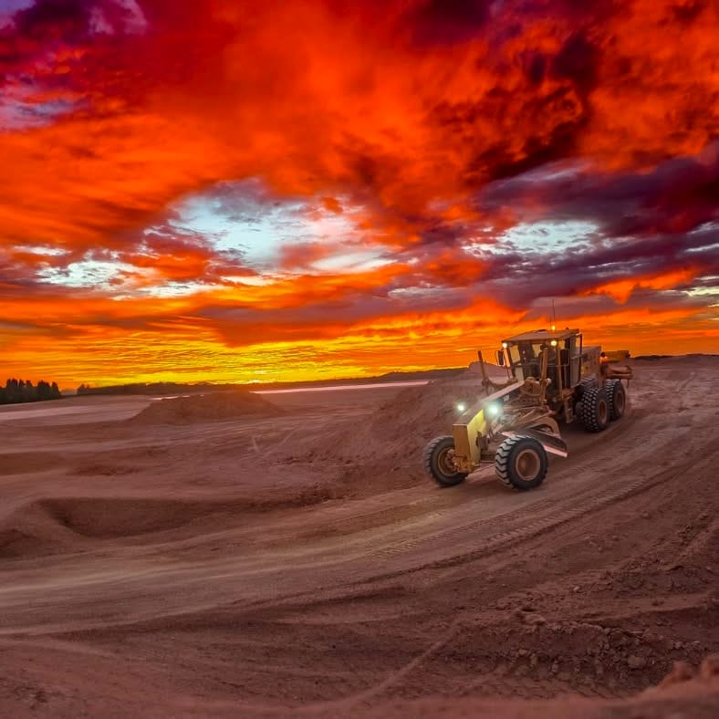 A grader cutting a red dirt haul road at last light, under a sky burning orange from horizon to horizon