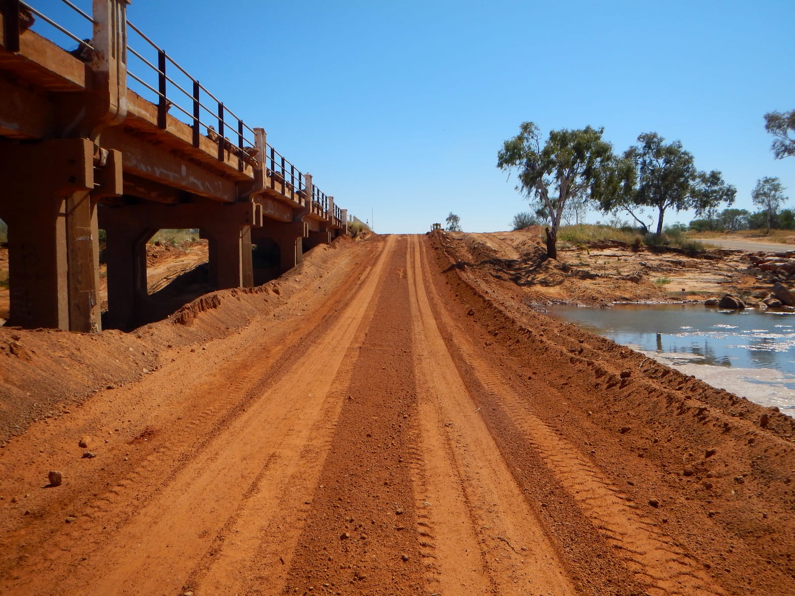 A newly resheeted red gravel road approaching a low concrete bridge, with a river pool visible alongside the embankment